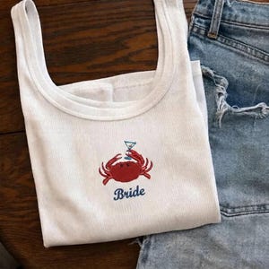 May include: A white ribbed vest top featuring an embroidered design of a red crab holding a blue martini glass. Below the crab, the word "Bride" is embroidered in blue script lettering. The garment is positioned next to blue denim fabric.