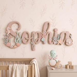 Könnte beinhalten: Ein hölzernes Wandschild mit dem Namen Sophia in Schreibschrift, verziert mit pastellfarbenen Meerestieren, Muscheln und Korallen. Darunter befinden sich eine hellgrüne Oktopus-Rassel, ein hölzernes Hasenspielzeug und drei kleine Buchstabenwürfel.