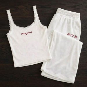 Op de afbeelding: Een witte tweedelige pyjamaset bestaande uit een hemdje met kanten rand en een bijpassende loungebroek. Het hemdje is voorzien van de tekst "mrs.jones" in donkerrood en de broek toont de datum "09.07.26" in dezelfde donkerrode kleur.