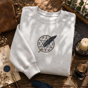 May include: A light gray crewneck sweatshirt featuring an embroidered design on the chest. The embroidery depicts a circular emblem with a six-pointed star, surrounded by symbols, and a dark blue quill pen placed diagonally across the center.
