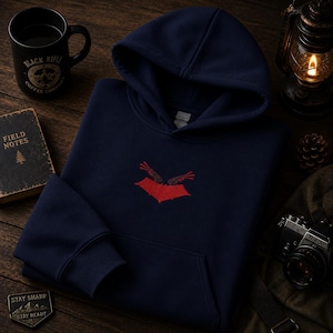 Peut inclure: Un sweat à capuche bleu marine avec un motif brodé rouge sur la poitrine. Le design représente deux armes de poing croisées au-dessus d'une forme rouge évoquant une chauve-souris. Le sweat possède des manches longues et une poche kangourou.