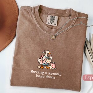 May include: An espresso-colored t-shirt featuring a graphic of a white goose wearing a pink apron and baking with a bowl and whisk. The text below the graphic reads, "Having a mental bake down." The shirt has a Comfort Colors brand label.