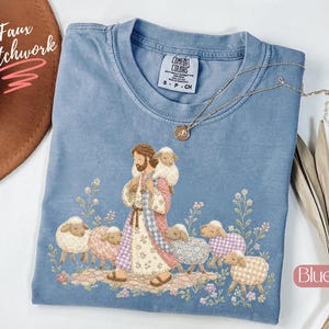 May include: A blue jean colored t-shirt featuring a graphic of a figure carrying a lamb on their shoulders, surrounded by several other sheep and floral elements. The shirt has a Comfort Colors label and a thin gold chain necklace rests on it.