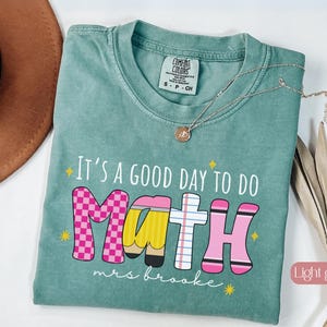 May include: A light green short-sleeve t-shirt with the text "It's a GOOD DAY TO DO MATH" and "mrs brooke" printed in white. The word "MATH" features letters styled as a pink checkerboard, a yellow pencil, lined paper, and a pink crayon.