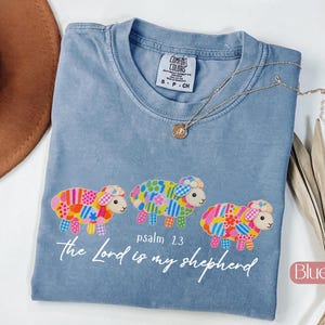 May include: A blue jean colored t-shirt featuring a graphic of three colorful patchwork sheep. Below the sheep, the text reads "psalm 23" and "the Lord is my shepherd" in white script. The shirt has a crew neckline and a Comfort Colors label.