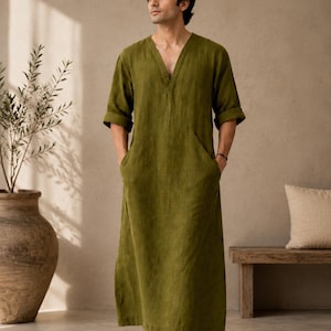 May include: An olive green ankle-length tunic with a V-neckline, short sleeves with folded cuffs, and two side pockets. The garment is made of a textured fabric and features a straight, loose-fitting silhouette that reaches down to the lower legs.