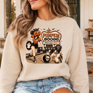Op de afbeelding: Een beige sweater met ronde hals en een grafische print van een persoon met een pompoenhoofd die aan het bowlen is. De tekst luidt PUMPKIN LANES, Strike if you DARE!, OPEN LATE en GHOULS NIGHT OUT. De illustratie bevat een bowlingbal met een schedel.