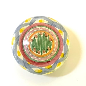 Peut inclure: Un bouton de meuble rond en céramique peint à la main. Il présente un motif central de feuilles vertes et de petits bourgeons bruns, entouré d'anneaux concentriques roses, oranges et verts. Le bord extérieur est orné d'un motif bleu et jaune.