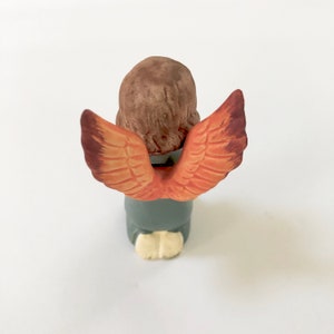 May include: A small ceramic angel figurine with brown hair, a gray robe, and orange wings. The angel is kneeling with its head bowed.