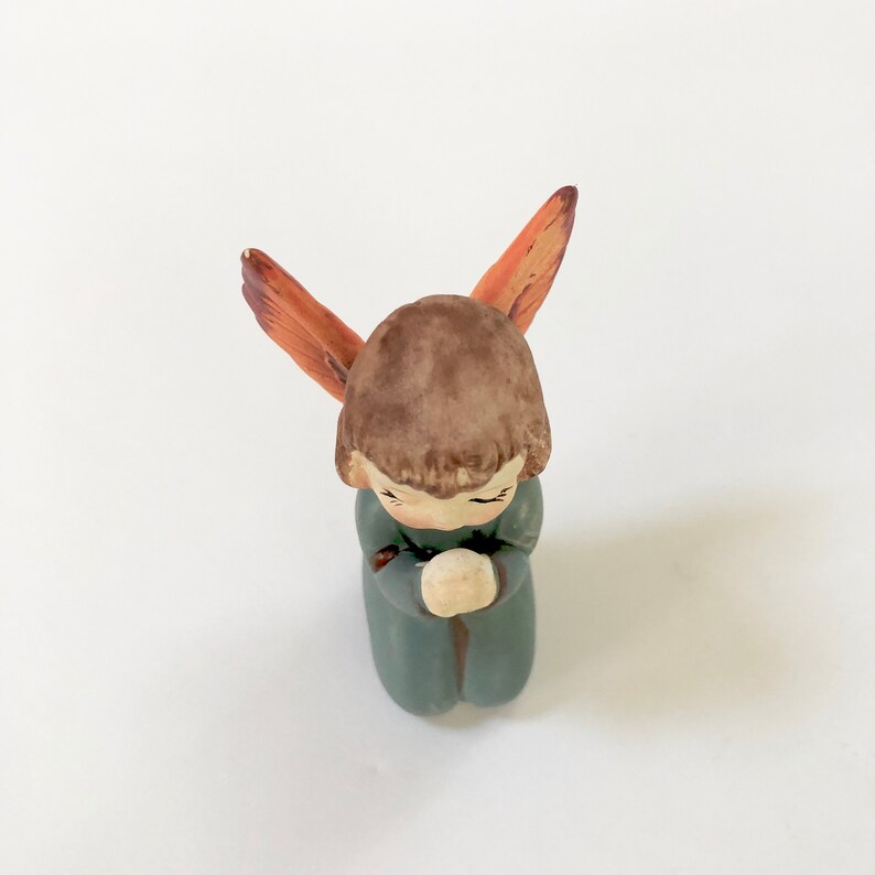May include: A small ceramic angel figurine with brown hair and wings, wearing a blue robe and praying with hands clasped.