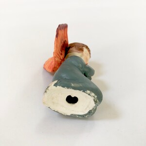 May include: A small ceramic figurine of an angel with orange wings, wearing a blue robe. The figurine is kneeling with its head bowed and has a hole in the bottom for hanging.