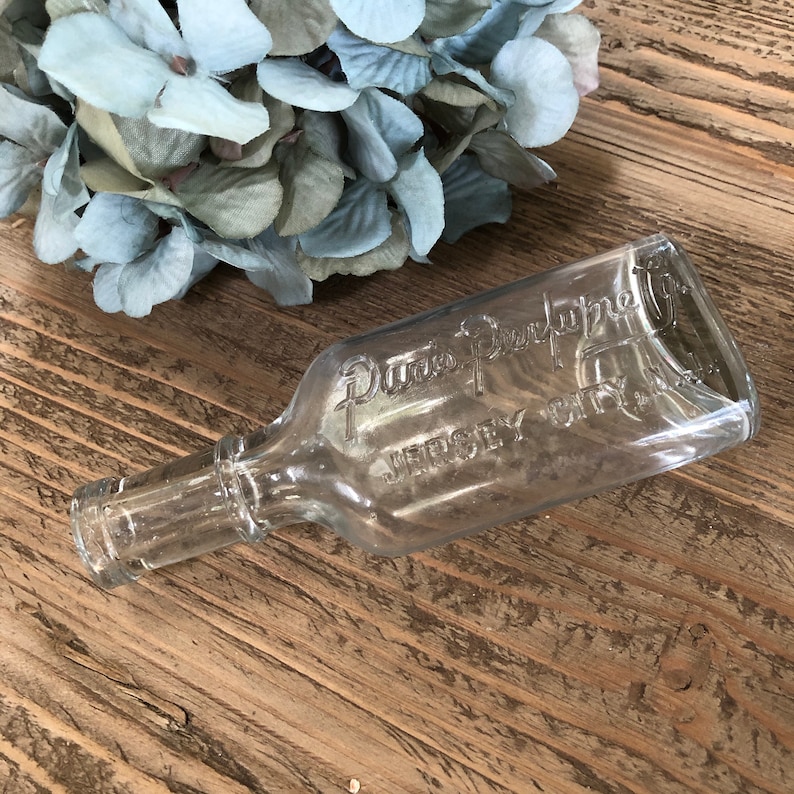 Puede incluir: Una botella de vidrio transparente con una superficie texturizada. La botella est&aacute; etiquetada con "Pure Perfumes" y "Jersey City, N.J."