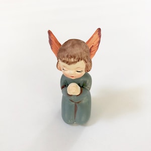 May include: A small, ceramic figurine of an angel kneeling with its hands clasped in prayer. The angel has brown hair, a blue dress, and orange wings.