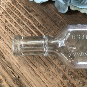 Puede incluir: Una botella de vidrio transparente con un cuello texturizado y una etiqueta que dice "S.H. 11".