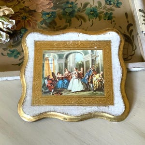 May include: A square decorative plaque with a scalloped edge, featuring a white distressed finish and a central gold frame. The centre displays a detailed scene of several figures in period clothing gathered within an architectural interior.