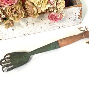 May include: A vintage garden hand rake with a wooden handle and a green metal head. The handle is a light brown colour, and the rake head has five tines. Dried flowers in a white wooden box are in the background.
