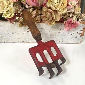 Pode incluir: Um ancinho de mão de jardim vintage com cabeça vermelha e cabo de madeira. O ancinho tem três dentes e é projetado para cultivar o solo. A alça é de cor marrom escuro. O ancinho está sobre um fundo de flores secas.