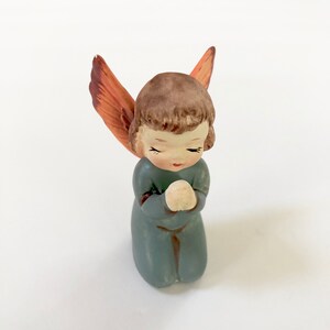May include: A small ceramic angel figurine with brown hair, closed eyes, and blue clothing. The angel is kneeling with hands clasped in prayer and has orange wings.