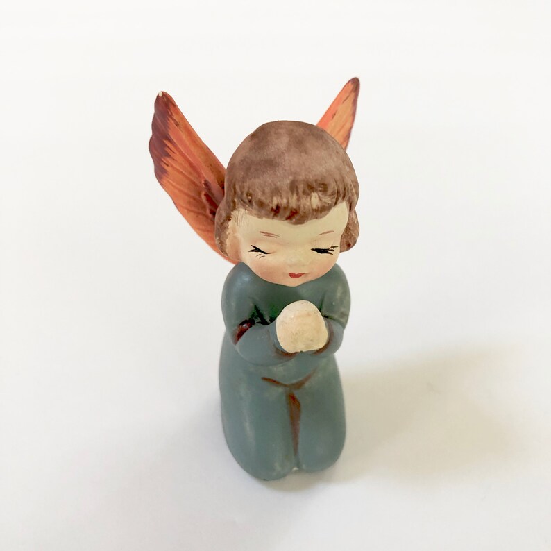 May include: A small ceramic angel figurine with brown hair, closed eyes, and blue clothing. The angel is kneeling with hands clasped in prayer and has orange wings.