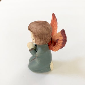 May include: A small ceramic figurine of an angel kneeling with its hands clasped in prayer. The angel has brown hair, a blue robe, and orange wings.