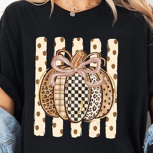 May include: A black short-sleeve t-shirt featuring a central graphic of a pumpkin with a bow. The pumpkin has segments with leopard print, plaid, and checkered patterns. Behind the pumpkin are vertical beige stripes with brown polka dots.