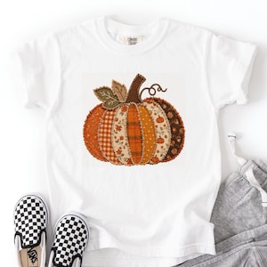 May include: A white short-sleeved t-shirt featuring a central graphic of a pumpkin composed of various patchwork fabric patterns in shades of orange, brown, and cream. The pumpkin design includes a brown stem and a single green leaf.