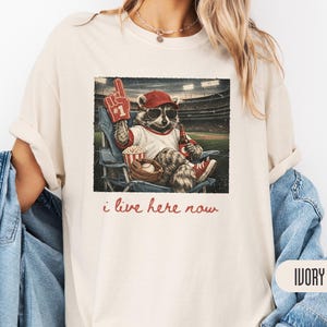 Op de afbeelding: Een ivoorkleurig t-shirt met een afbeelding van een wasbeer met een rode honkbalpet en zonnebril, zittend in een stadionstoel met een schuimvinger, popcorn en een drankje. De tekst onder de afbeelding luidt, i live here now.