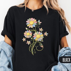 May include: A black short-sleeved t-shirt featuring a central graphic of three large flowers and smaller star-shaped blossoms. The petals are composed of various colourful plaid and gingham patterns in shades of yellow, pink, and blue.