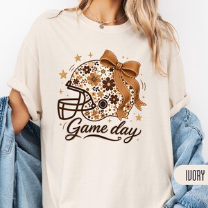 Może przedstawiać: T-shirt z krótkim rękawem w kolorze kości słoniowej z grafiką kasku do futbolu amerykańskiego ozdobionego brązowymi i beżowymi wzorami kwiatowymi oraz dużą brązową kokardą. Napis "Game day" widnieje poniżej ciemnobrązową kursywą.