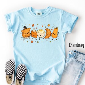 May include: A light blue t-shirt featuring a graphic of five cartoon food items including a turkey, a slice of pie, a bowl of mashed potatoes, a croissant, and a squash, all with smiling faces, surrounded by small autumn leaves and hearts.