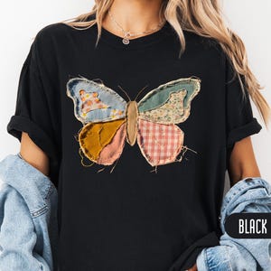 May include: A black t-shirt featuring a large butterfly graphic on the front. The butterfly wings are composed of various fabric patches in blue, yellow, pink, and white with floral and gingham patterns. The word BLACK is printed in white text.
