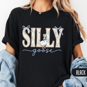 Puede incluir: Una camiseta negra con un estampado gráfico que muestra el texto SILLY en un patrón floral y de cuadros, acentuado con lazos azul claro. La palabra goose está escrita debajo en cursiva, junto a una ilustración de un ganso con un gorro.