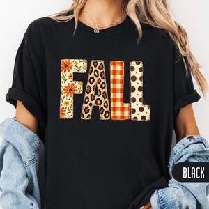 Op de afbeelding: Een zwart T-shirt met korte mouwen en het woord FALL in grote blokletters. Elke letter heeft een eigen patroon: de F heeft oranje bloemen, de A een luipaardprint, de L een oranje ruitpatroon en de tweede L heeft bruine stippen.