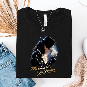 May include: A black crew neck t-shirt featuring a graphic print of Michael Jackson wearing a black top hat and a sequined jacket, with the name Michael Jackson written in gold cursive script below the image and a silver sunburst pendant necklace.