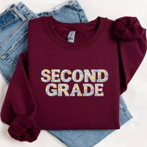 May include: A maroon crewneck sweatshirt featuring the text SECOND GRADE in a multi-coloured pastel patchwork pattern. The letters have a grid-like texture with various soft floral and geometric designs in shades of pink, blue, yellow, and green.