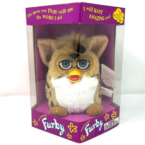 May include: A vintage Electronic Furby toy in its original packaging. The Furby has a tan and brown spotted pattern, large blue eyes, and a red beak. The packaging is purple and white with the Furby logo and text that reads "The more you PLAY with me the MORE I do!"