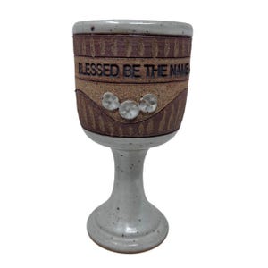 May include: A ceramic chalice with a wide, textured brown body and a light gray stem and base. The chalice features the phrase "BLESSED BE THE NAME" in black lettering and three decorative white accents.