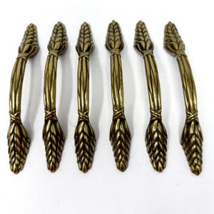 May include: A set of six antique brass-toned metal cabinet pulls, each featuring a textured, elongated wheat stalk design with a central band detail. The handles have a curved, arched shape with detailed, layered leaf-like patterns at both ends.