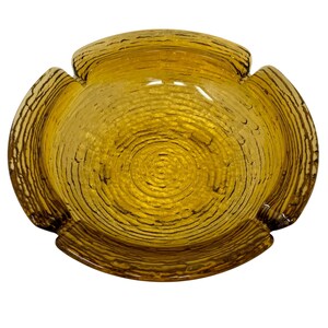 May include: A vintage amber-colored glass ashtray with a textured, ridged surface. The ashtray has a four-lobed design with a circular interior and a series of concentric circles. The glass has a warm, golden hue, and the ridges create a pattern.
