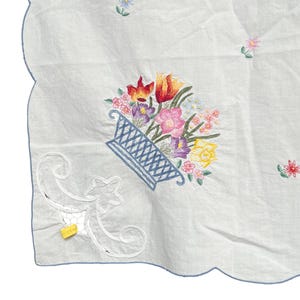May include: White embroidered cloth with a scalloped blue edge. A blue basket filled with colorful flowers is embroidered in the center. Additional small floral embroidery is scattered across the cloth.