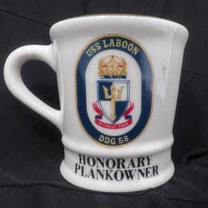 May include: White ceramic mug with a navy blue and gold crest featuring a trident and the text "USS LABOON DDG 58 WITHOUT FEAR". The bottom of the mug reads "HONORARY PLANKOWNER".