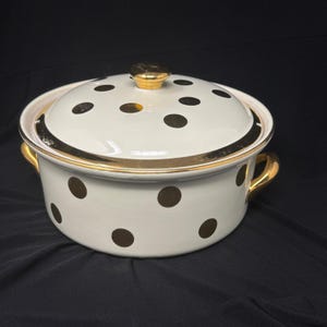 May include: Cream-colored enamelware pot with a lid, adorned with dark brown polka dots. The pot features gold-colored handles and trim, including the lid's knob. The pot is designed for cooking and serving.