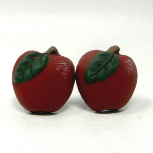 May include: Two red apple-shaped decorative objects with green leaves and brown stems. The apples have a textured surface and are set against a white background. These could be used as decorative accents.
