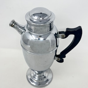 May include: A silver cocktail shaker with a black handle. The shaker has a rounded body and a tall, slender spout. The handle is attached to the side of the shaker with a hinge.