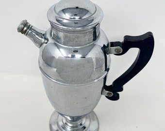 Op de afbeelding: Een zilveren cocktailshaker met een zwarte handgreep. De shaker heeft een rond lichaam en een lange, slanke tuit. De handgreep is met een scharnier aan de zijkant van de shaker bevestigd.