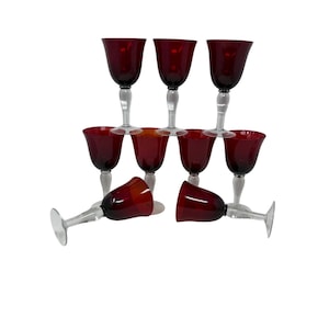 May include: A collection of nine wine glasses with ruby red bowls and clear stems and bases. The glasses are arranged in a pyramid shape, with some lying on their sides. The background is plain white.