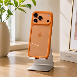 May include: A smartphone with a translucent orange protective case featuring a raised camera frame, resting on a silver metal stand. The phone displays a clear back panel with a visible Apple logo and a triple-lens camera system on the upper left.