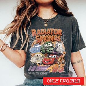 May include: A charcoal grey short-sleeve t-shirt featuring a graphic print of characters from Radiator Springs dressed in Halloween costumes. The text reads Radiator Springs and Trunk or Treat with the number 95. The design includes spiderwebs.