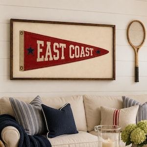 May include: A framed red felt pennant with the text EAST COAST in white lettering, featuring two dark blue stars. The pennant is displayed on a white wall above a beige sofa with blue striped and solid navy pillows, a navy throw, and a wooden coffee table.