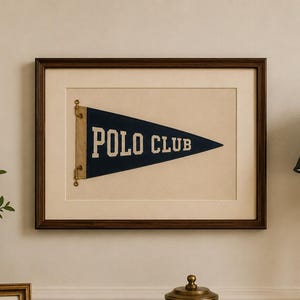 Op de afbeelding: Een ingelijste marineblauwe driehoekige wimpel met de tekst POLO CLUB in witte letters, gemonteerd op een cr&egrave;mekleurige passe-partout in een donkerhouten lijst. De wimpel is aan de linkerzijde bevestigd met messing beslag en toont een sportief ontwerp.